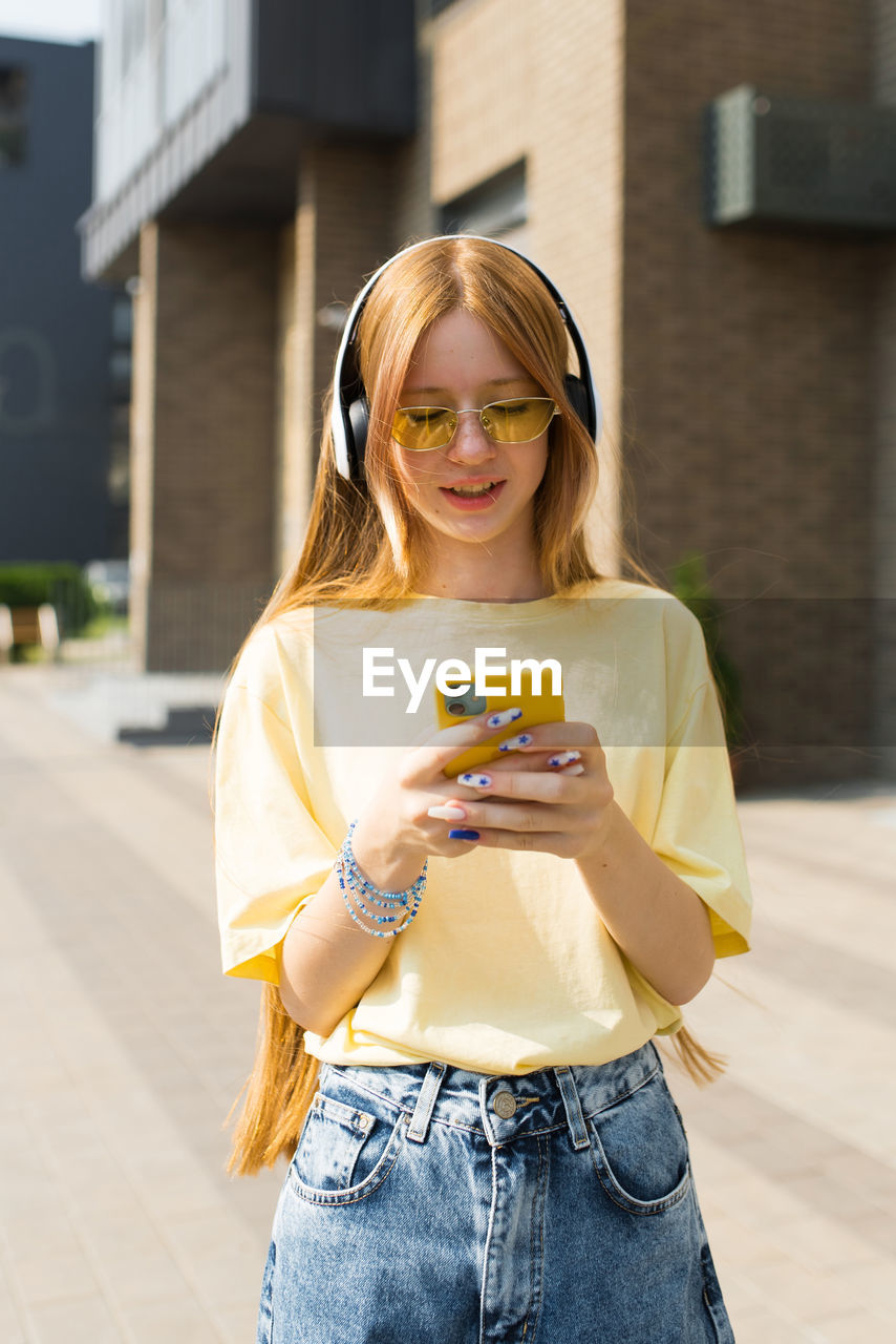 Caucasian teenager girl in a yellow shirt is looking at her cell phone while wearing headphones