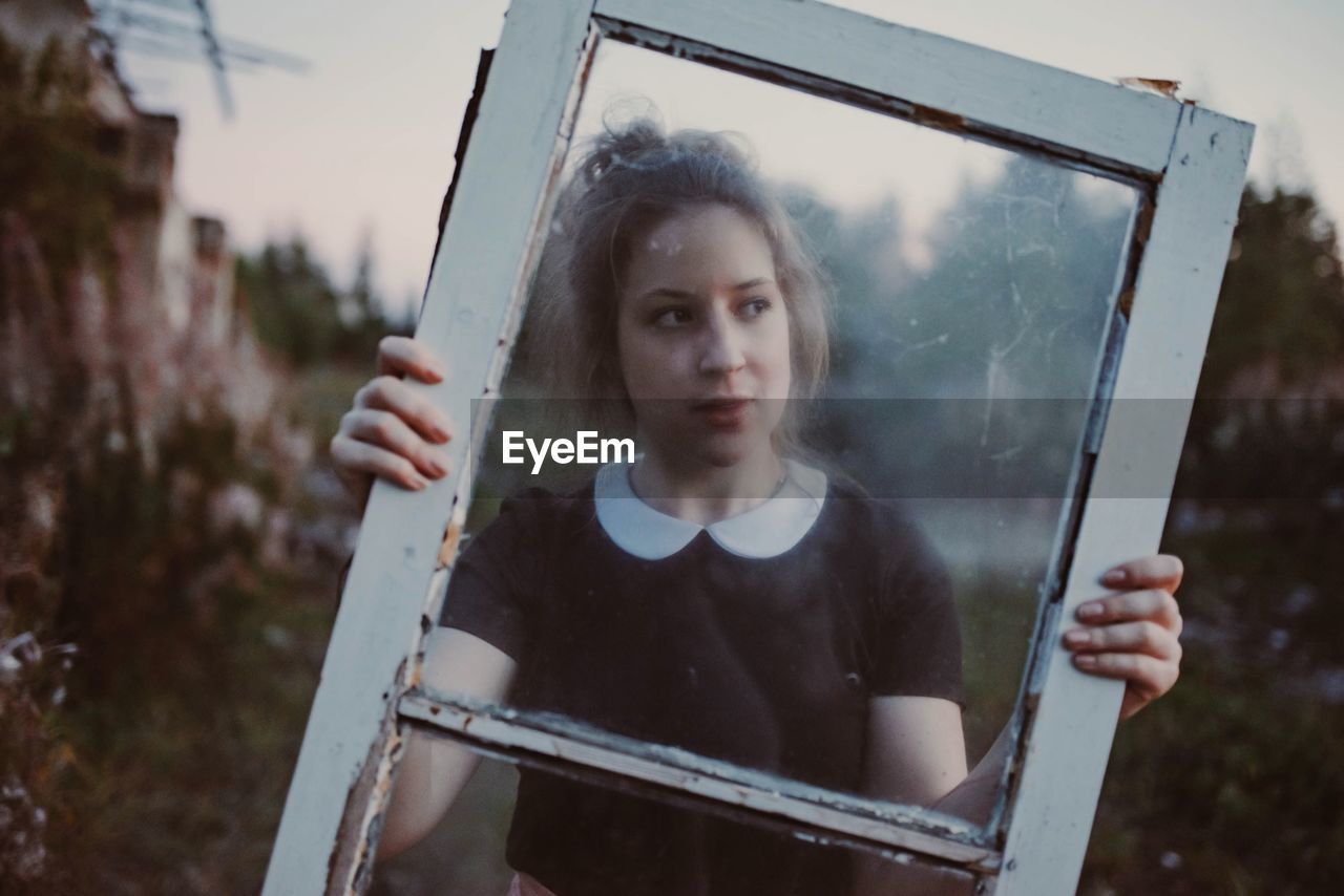 Portrait of young woman holding window while standing outdoors