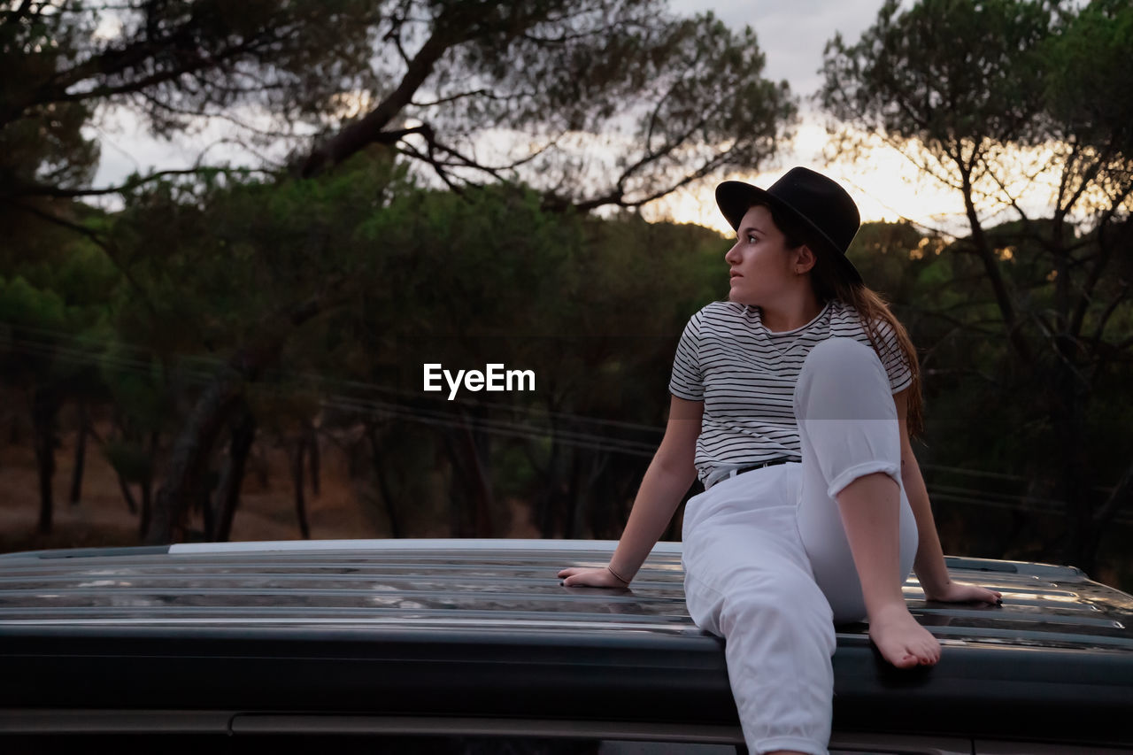 Young woman looking away while sitting on railing against trees