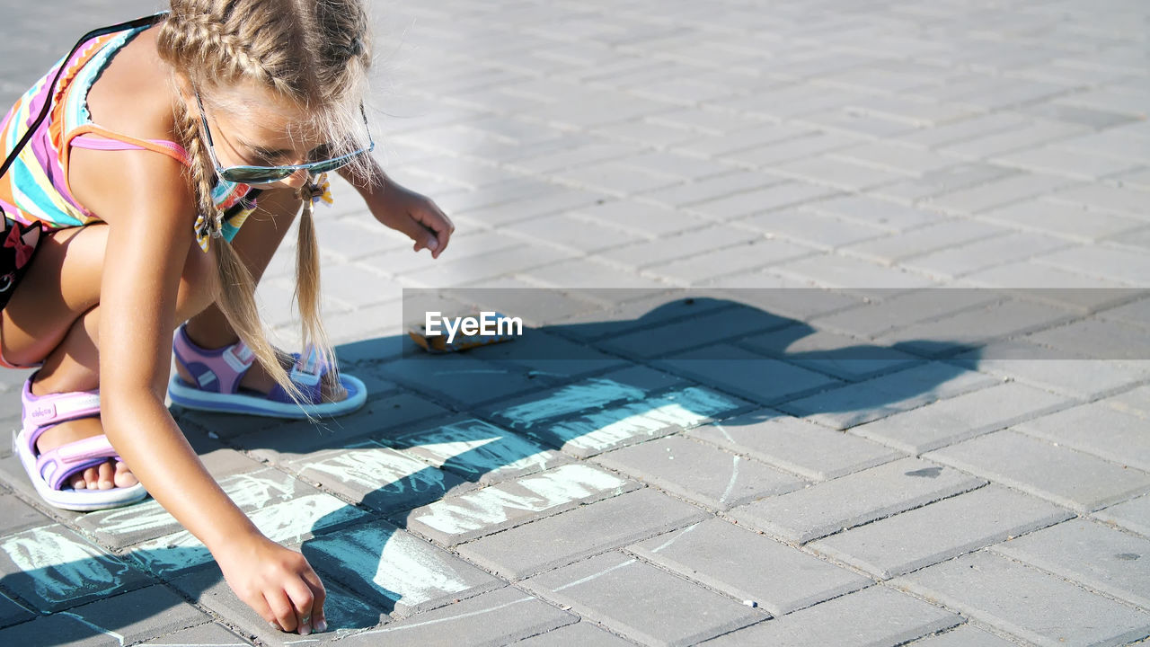 A girl in sunglasses, draws drawings with colored crayons on the asphalt, street tiles. a hot