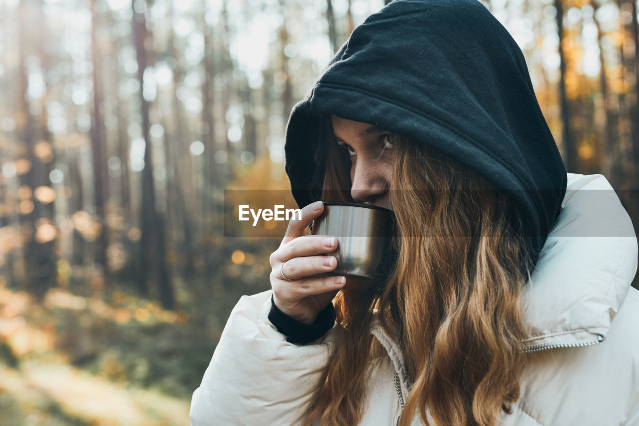 Woman in a hood having break during autumn trip holding cup with hot drink from thermos flask