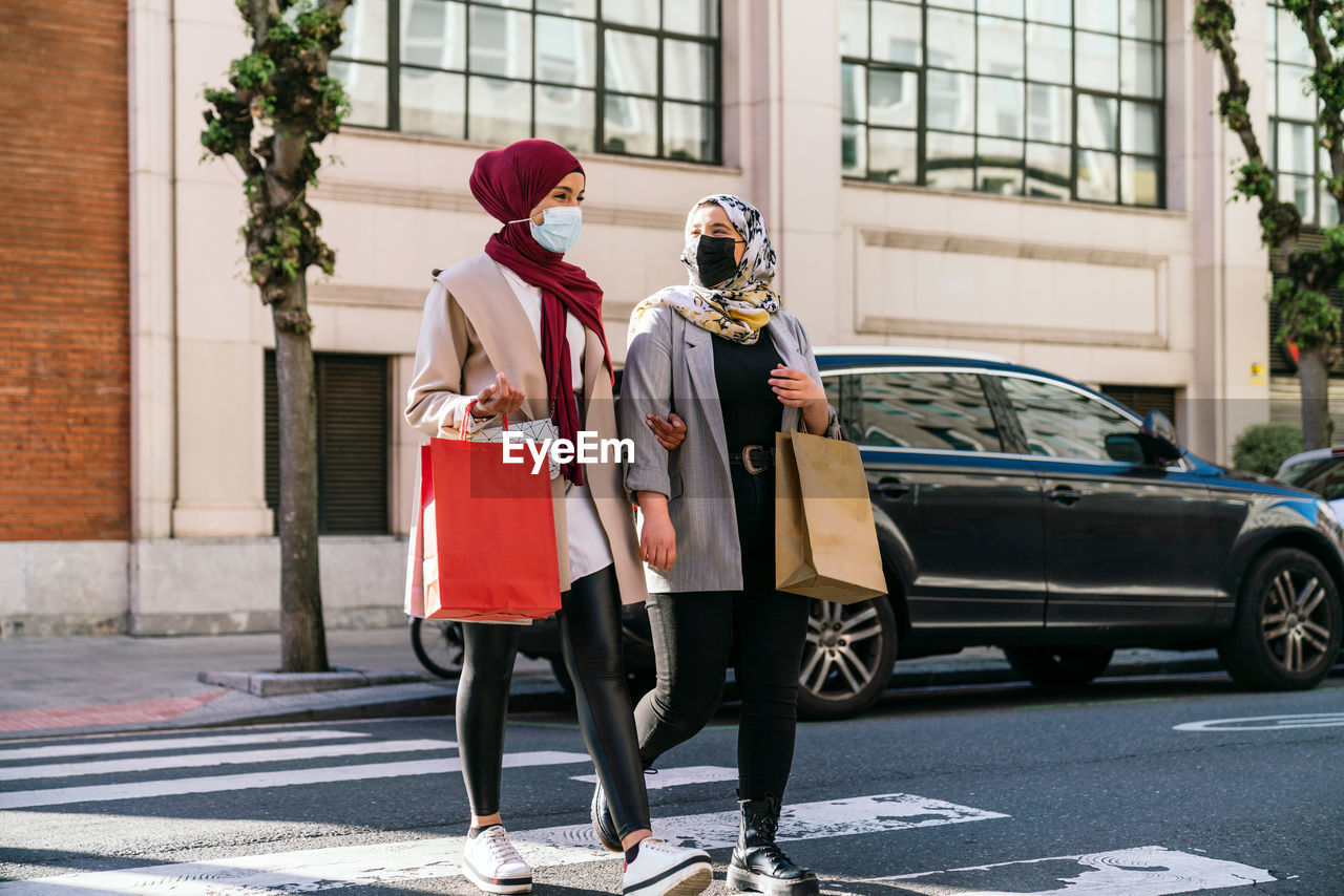Muslim female friends in masks and with paper bags crossing road while walking in city after shopping