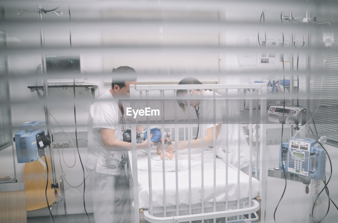 Faceless doctors standing near hospital bed with newborn in intensive care unit