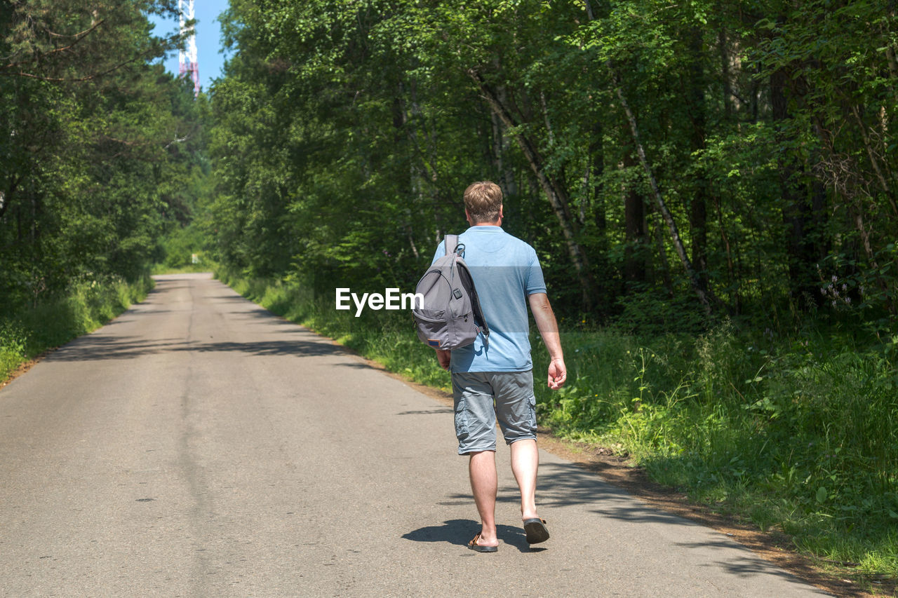 An adult male traveler with a backpack is walking alone empty road in the middle forest.