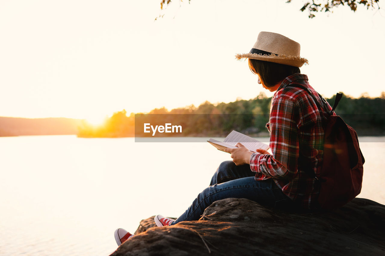 SIDE VIEW OF MAN SITTING ON BOOK BY LAKE AGAINST SKY