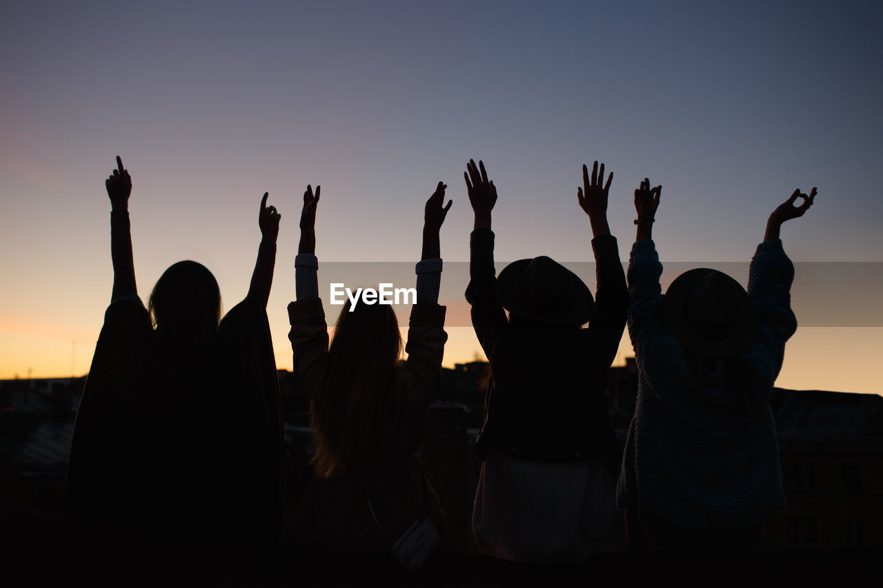 Silhouette photo of four attractive happy young women sitting with their backs and hands up.