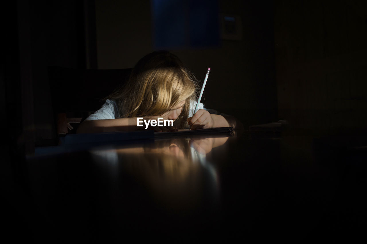 Girl writing homework while sitting at table in darkroom in house