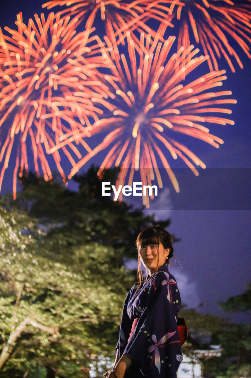 Low angle view of young woman standing against firework display at dusk