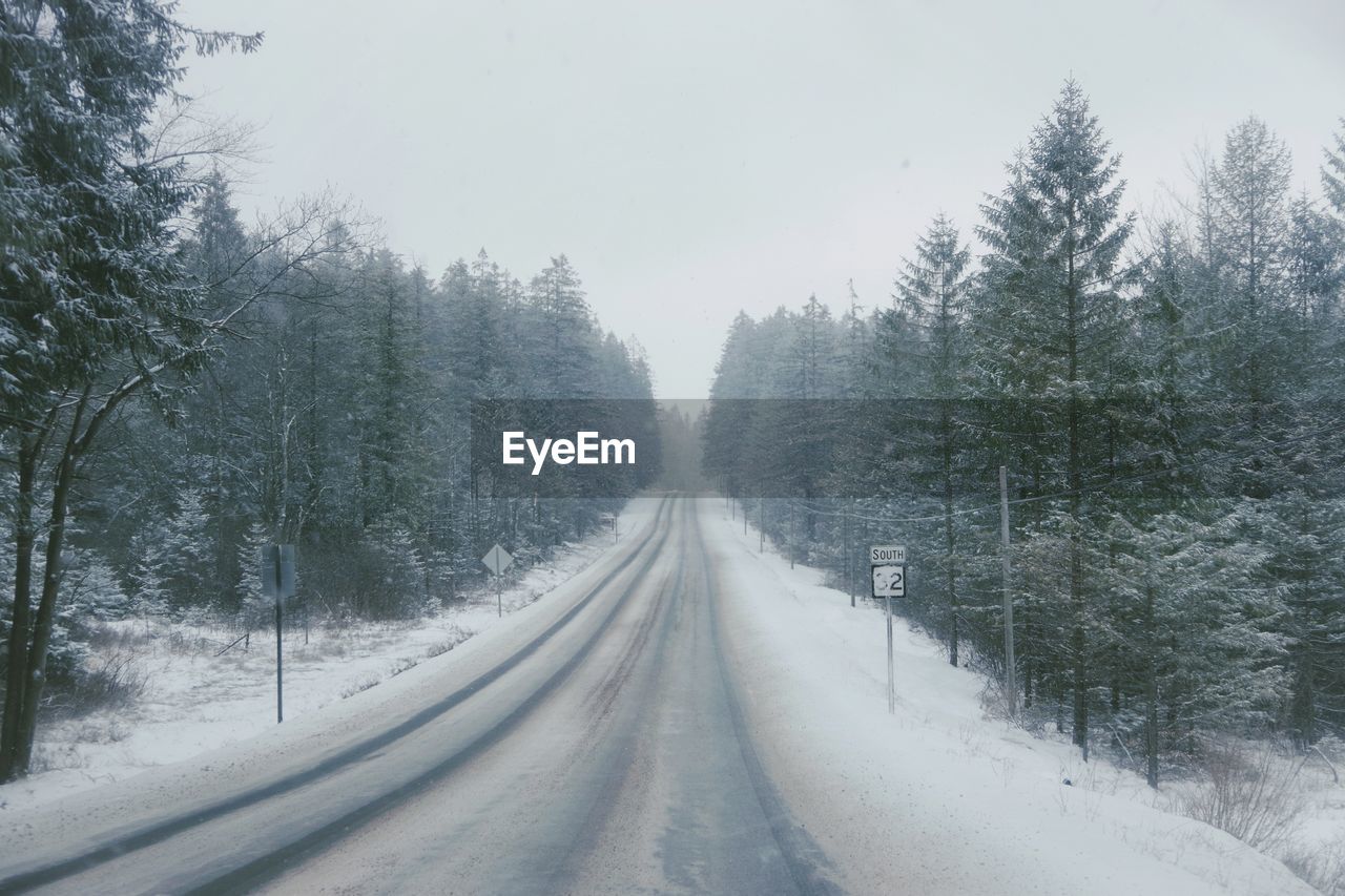 Snow covered road amidst trees against sky in winter