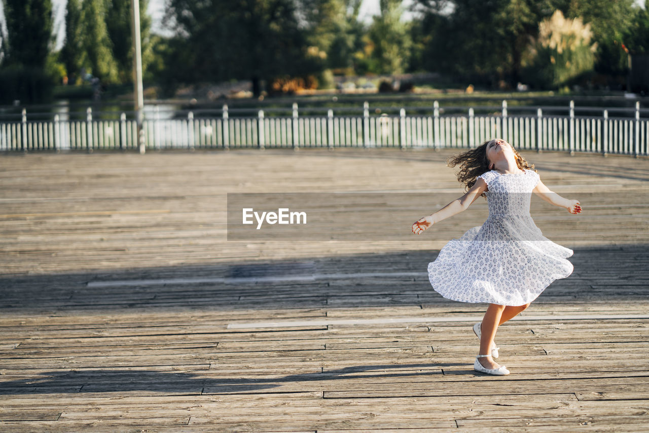 Cute teen girl spinning with hands apart on boardwalk of embankment