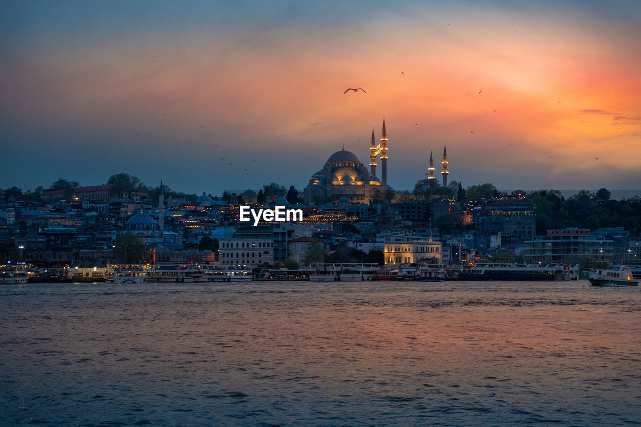 buildings by river against sky at sunset