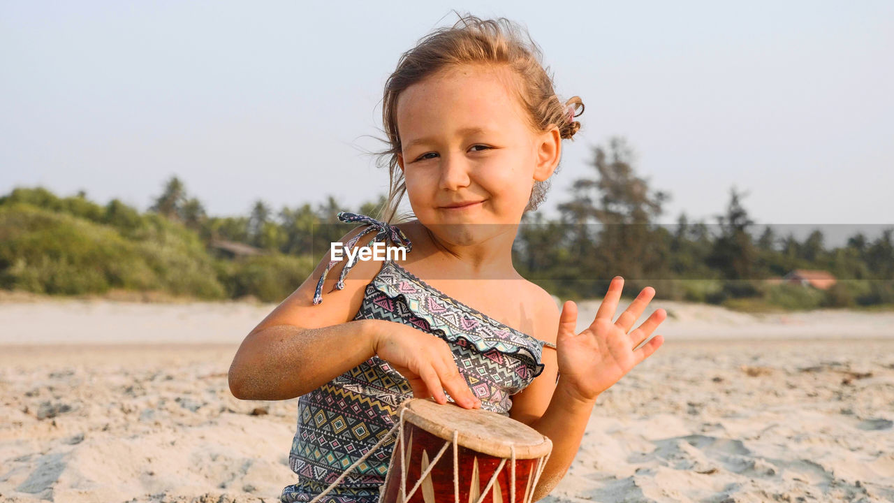 Portrait of smiling girl playing drum while sitting at beach