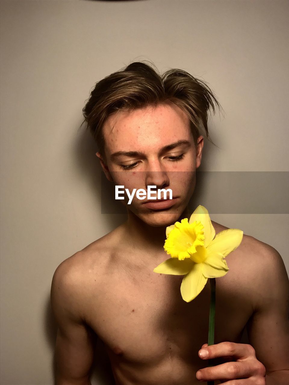 Shirtless young man holding yellow daffodil against wall