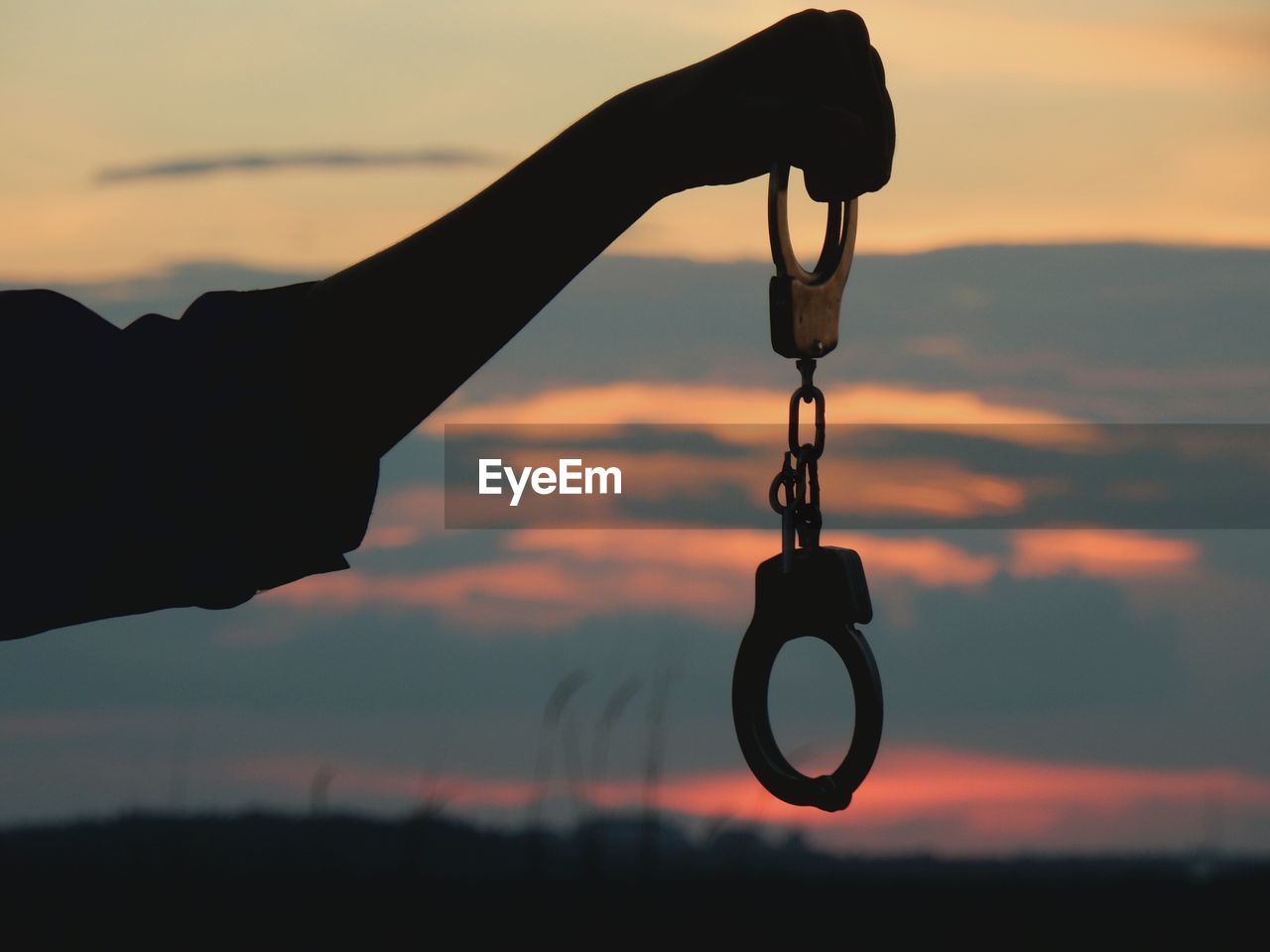Close-up of silhouette hand holding handcuffs against sky during sunset