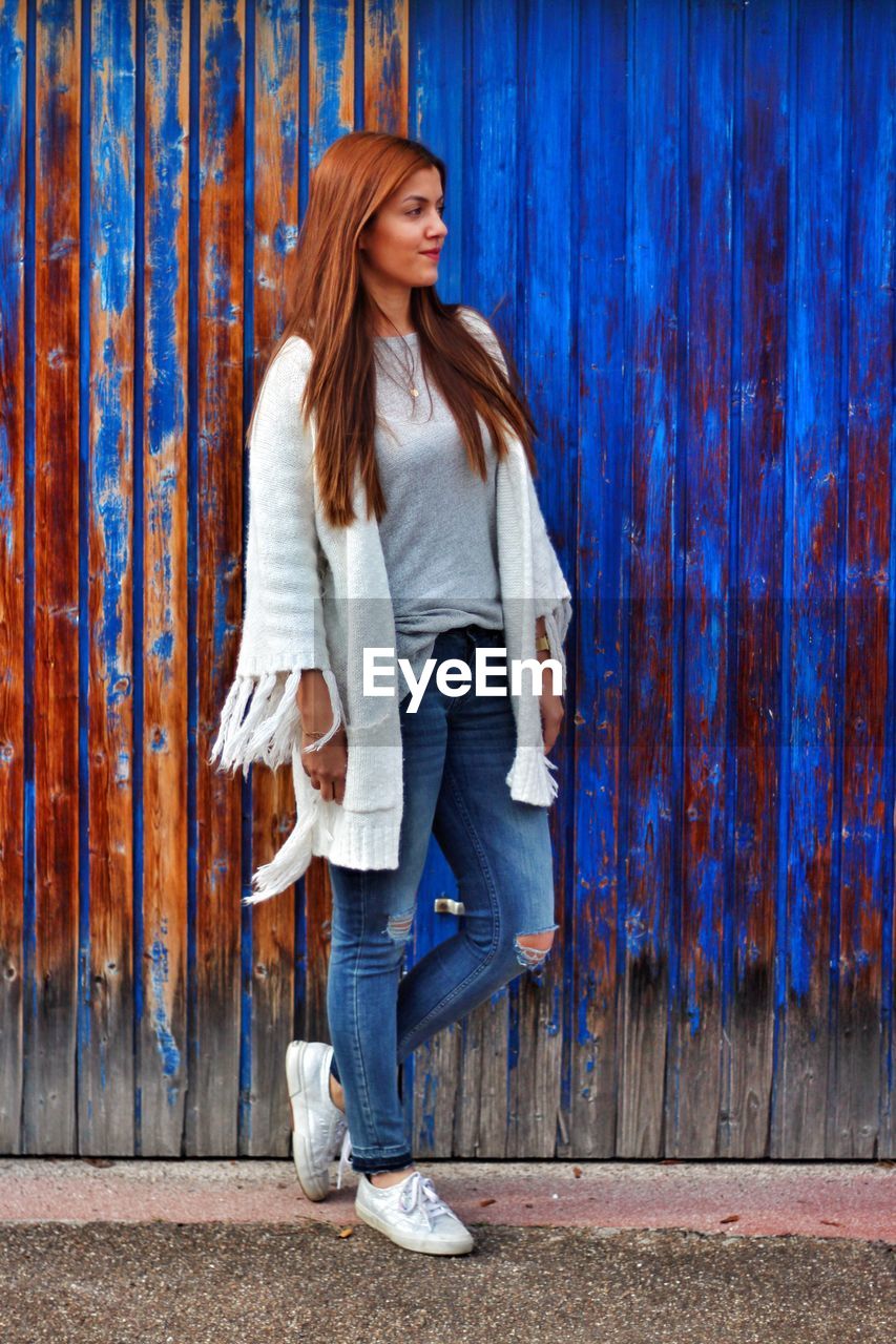 Full length of young woman looking away while standing against blue weathered wall