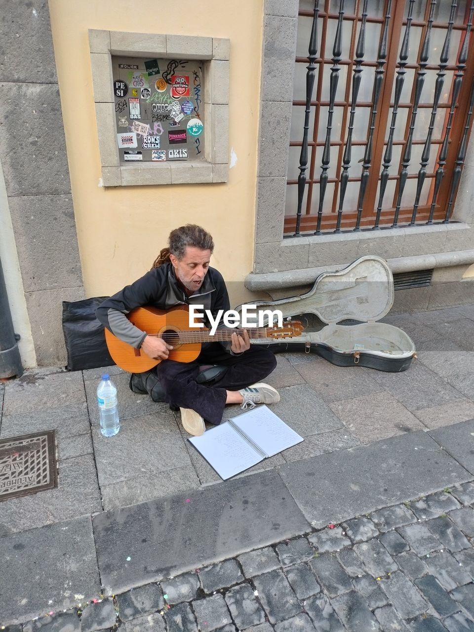 string instrument, guitar, musical instrument, music, musician, one person, full length, sitting, adult, arts culture and entertainment, musical equipment, street artist, plucking an instrument, men, street musician, day, leisure activity, city, young adult, architecture, holding, front view, casual clothing, acoustic guitar, performance, person, lifestyles, high angle view, creativity, electric guitar, wall, footpath, street, outdoors