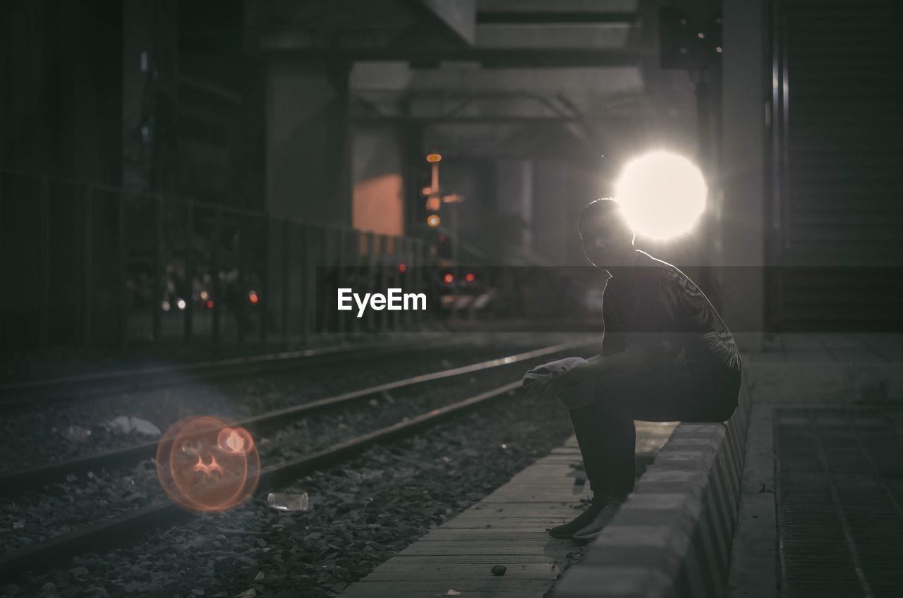 Side view of man sitting by railroad tracks at night