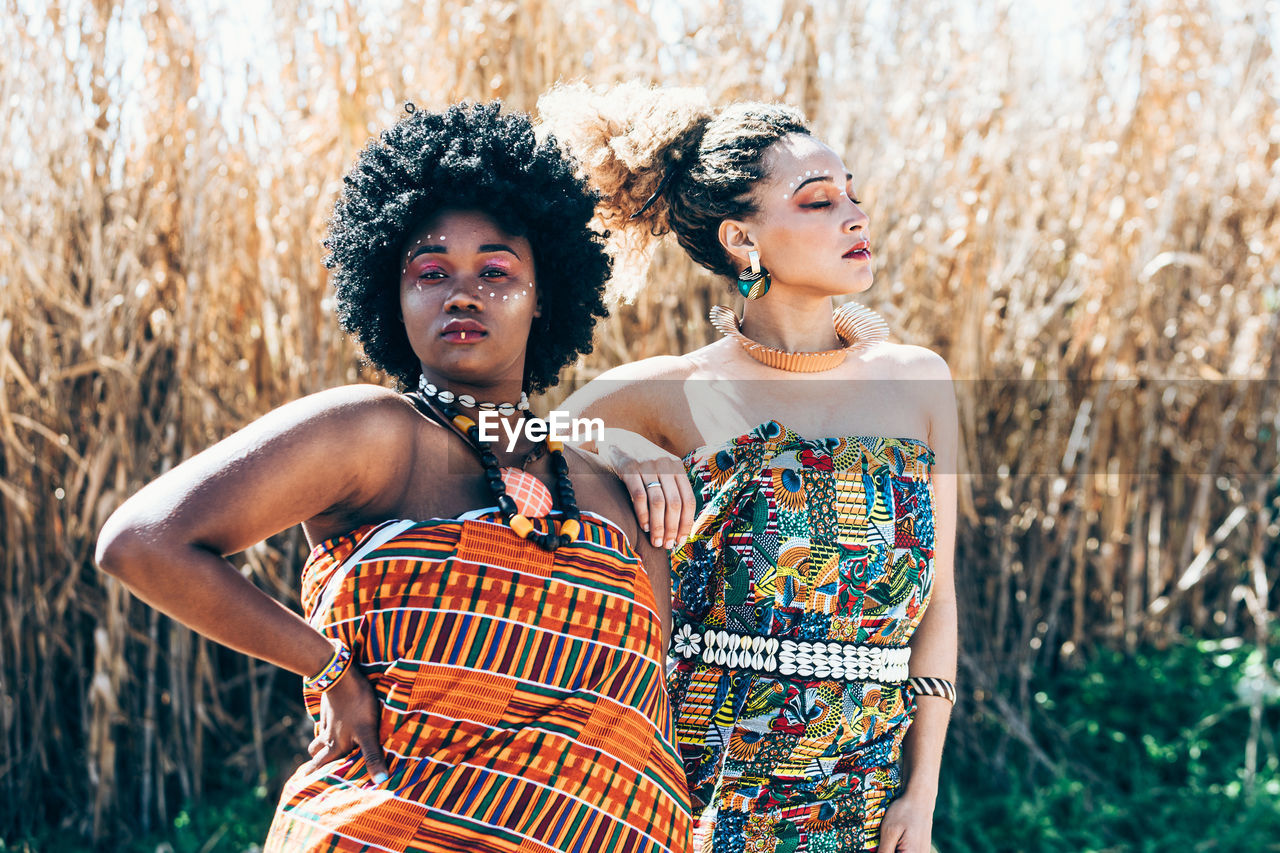 Confident black and mixed race female models in ornamental dresses with authentic african accessories looking at camera against dry grass in sunlit field