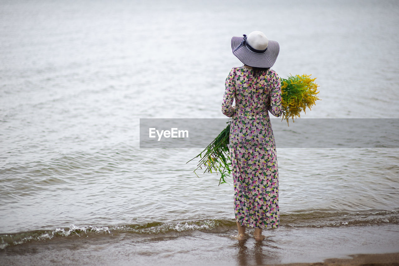 Rear view of a slide woman with a bouquet of wild flowers standing on the sea shore