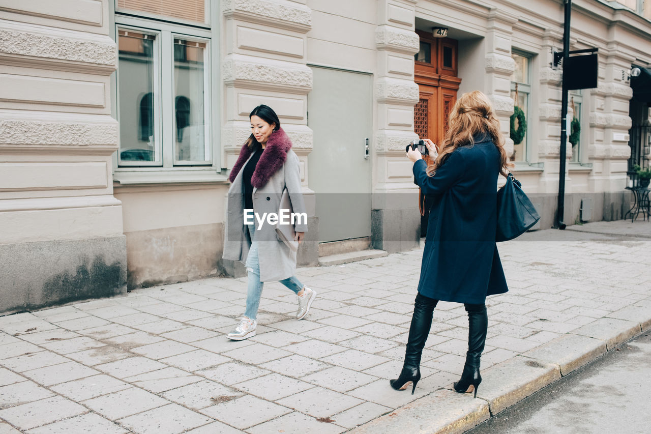 Rear view of woman photographing female friend walking on sidewalk against building in city