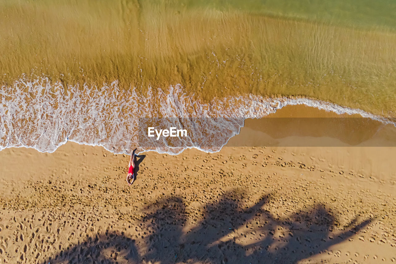 Woman lying on the beautiful sandy beach touching ocean waves with her feet, drone aerial view.