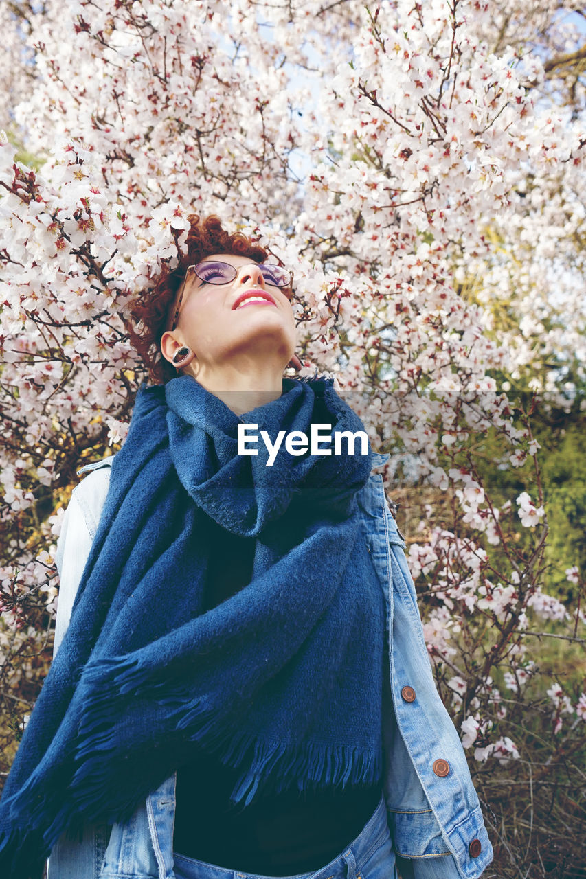 Beautiful young woman looking up while standing against cherry tree