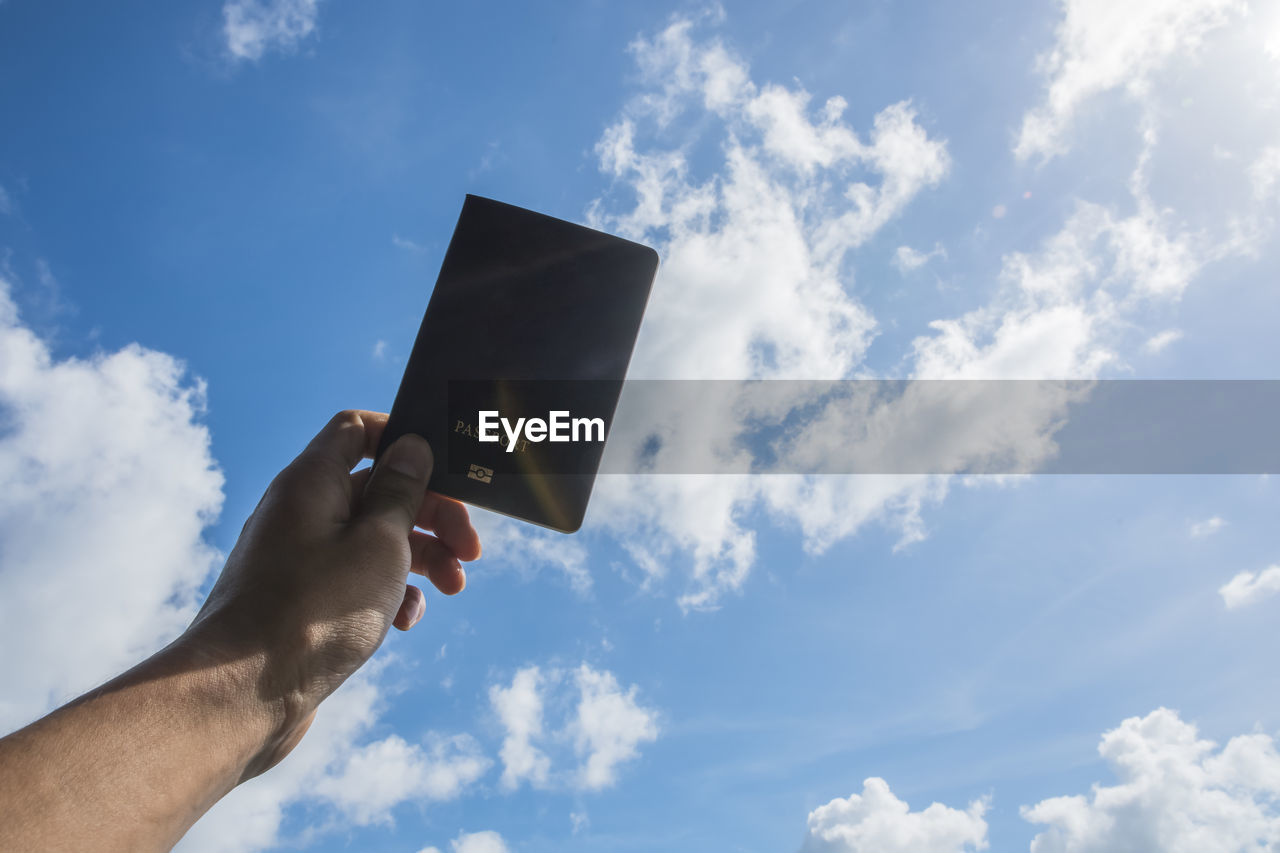 Cropped image of person holding passport against sky