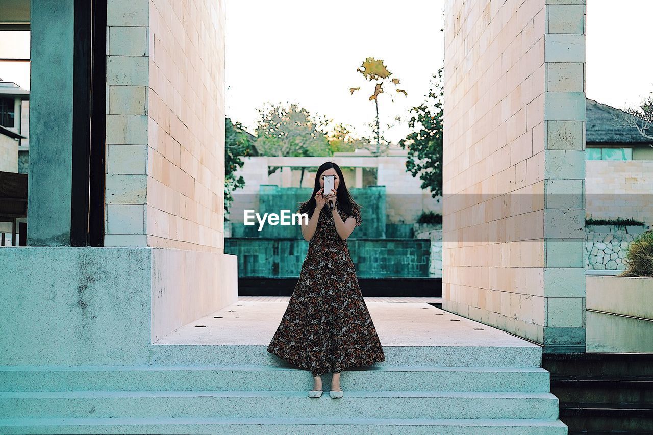 YOUNG WOMAN STANDING AGAINST BUILT STRUCTURE