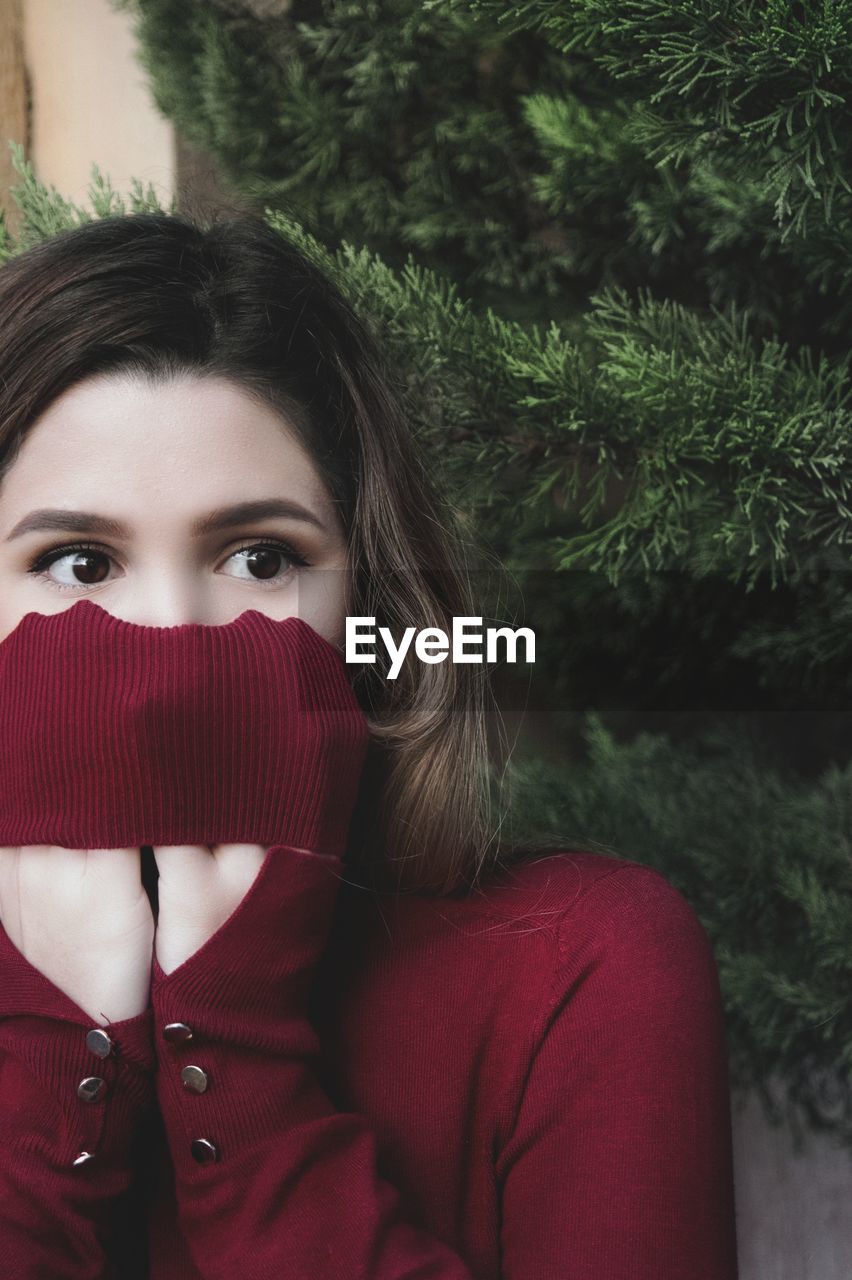 Close-up of shocked woman covering face with sweater against tree