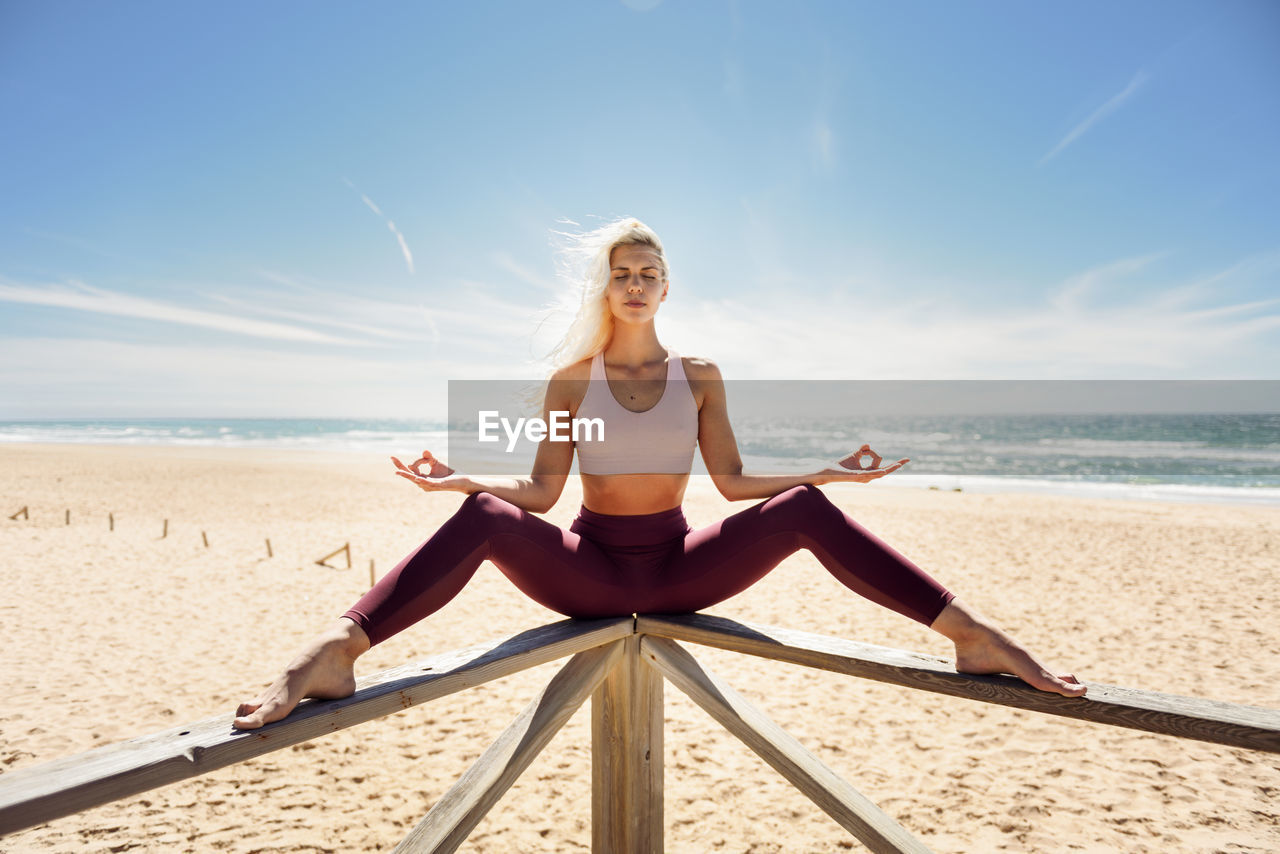 Full length of young woman doing yoga on railing at beach during sunny day