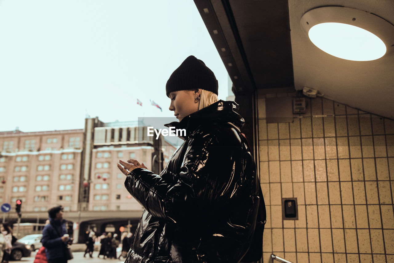 Side view of young woman wearing black shiny jacket while standing at subway's entrance in city during winter