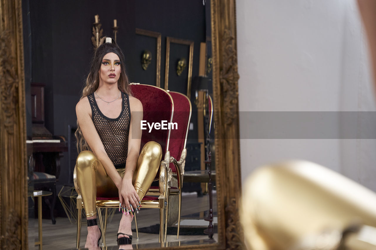 Reflection of non binary person wearing golden leggings sitting on chair at home