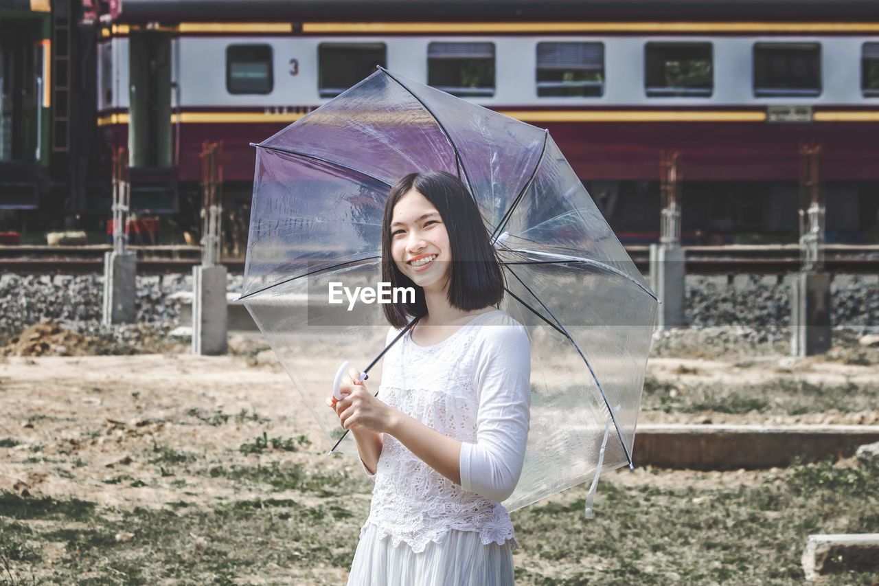 Portrait of smiling young beautiful women holding umbrella standing in front of train.