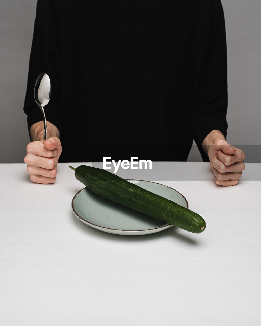 Midsection of person holding spoon with cucumber in plate on table