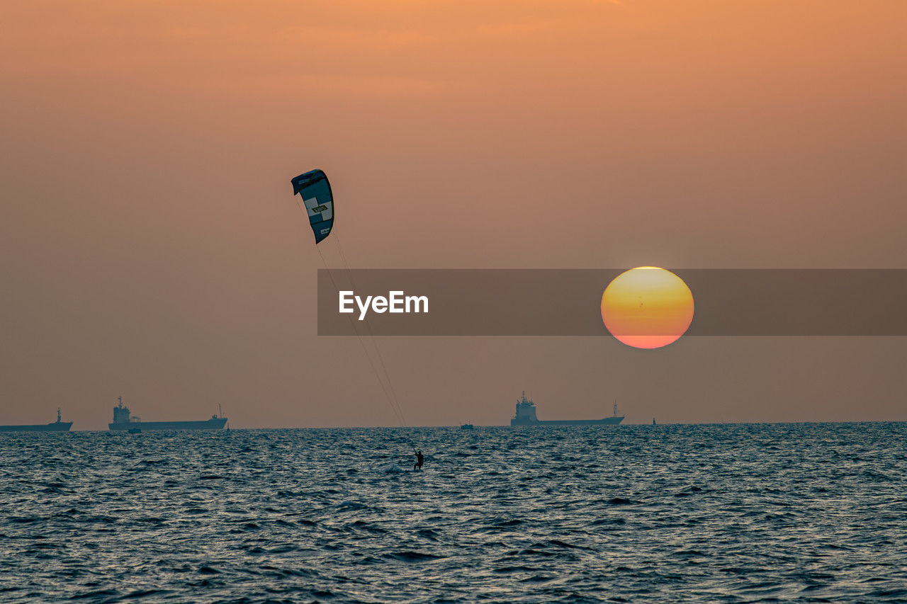 sky, sea, water, sunset, horizon, ocean, nature, beauty in nature, transportation, adventure, scenics - nature, horizon over water, sports, flying, sun, tranquility, nautical vessel, tranquil scene, travel, orange color, parachute, outdoors, environment, motion, balloon, travel destinations, silhouette, windsports, sunlight, mid-air, wave, vacation, extreme sports, idyllic, mode of transportation, holiday, trip, coast, rippled, dusk, leisure activity, waterfront, land, urban skyline, kite sports, dramatic sky, no people