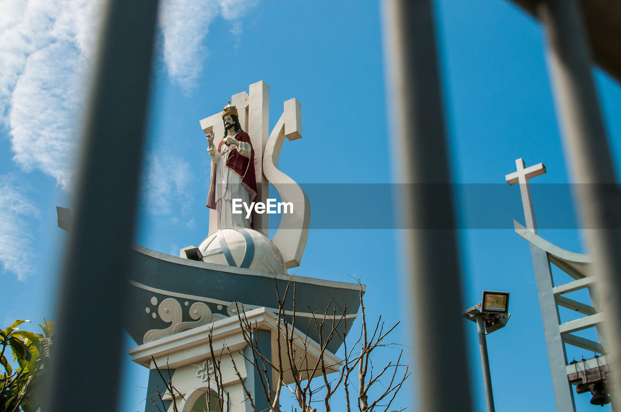 Low angle view of jesus christ against sky seen through fence