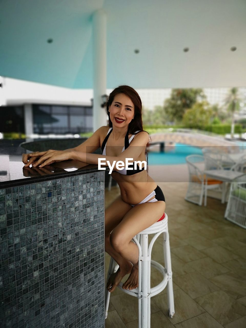 Portrait of woman wearing bikini while sitting on stool at poolside