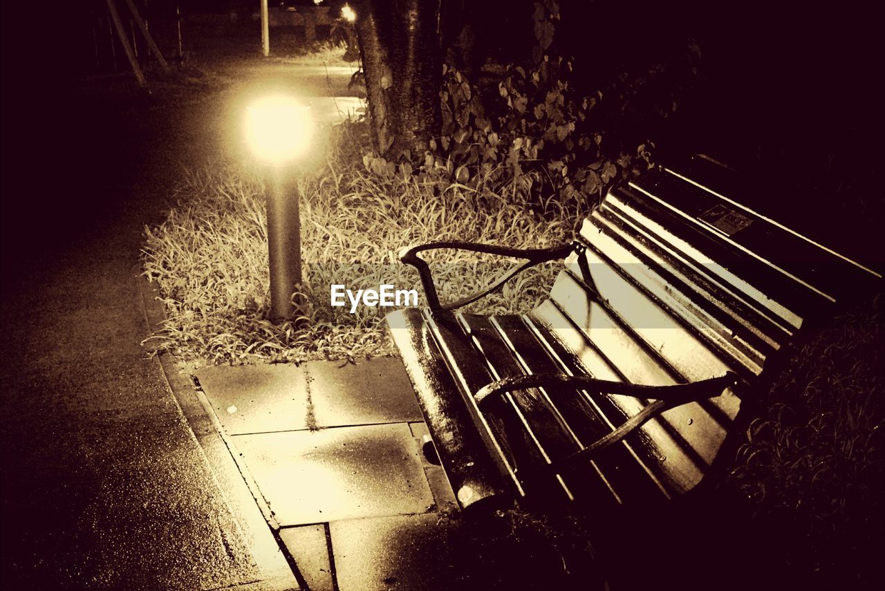 Empty wooden bench by illuminated bollard in garden at night