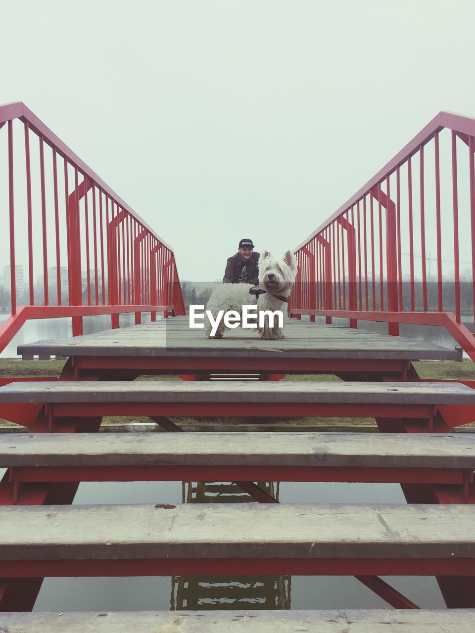 Dog with young man in background standing on footbridge against sky