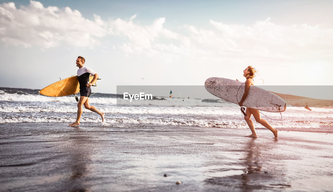 Cheerful couple running on beach with surfboards