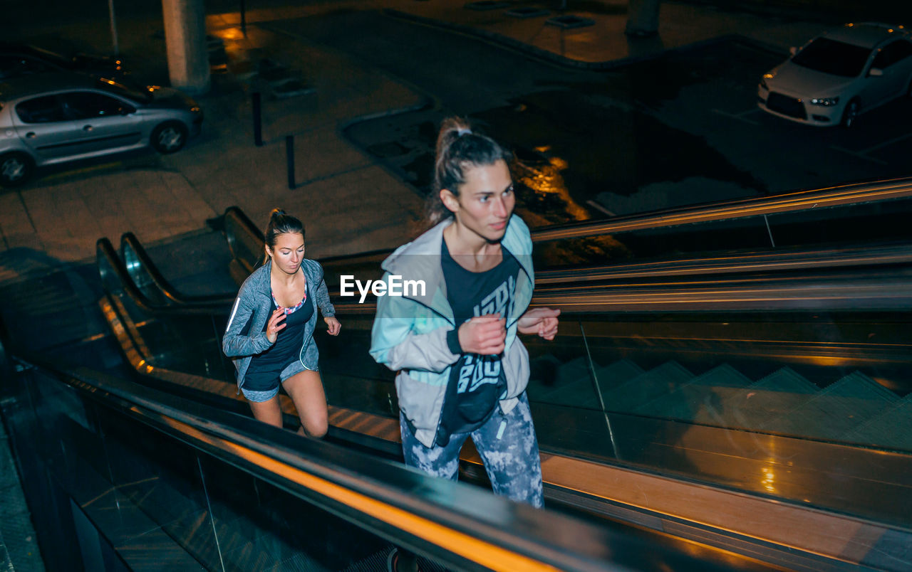 Women friends training running up escalator in city at night