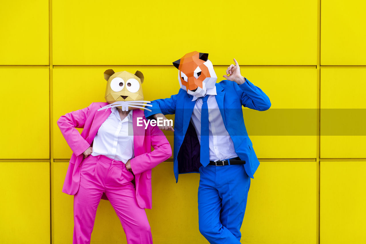 Man and woman wearing vibrant suits and animal masks posing together against yellow wall