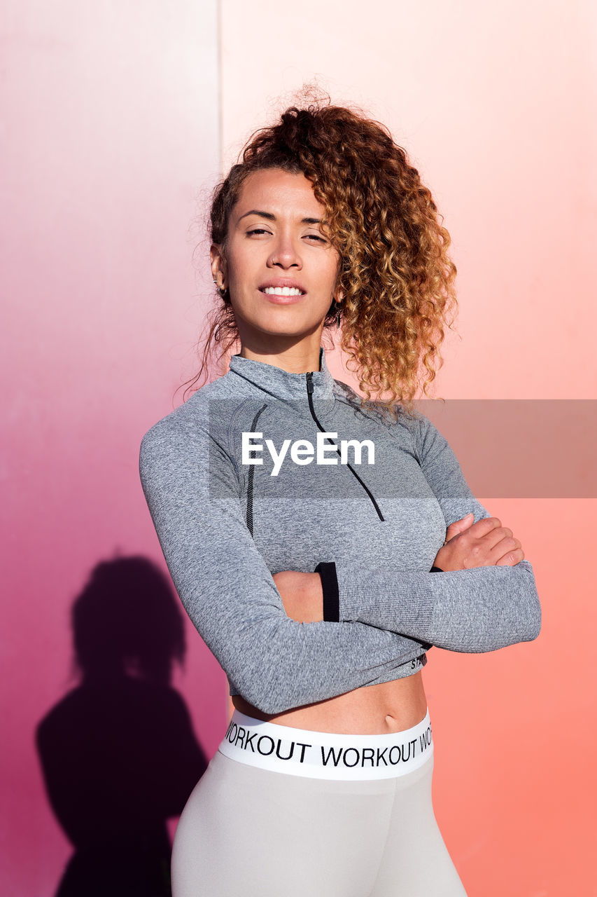 Self confident adult female athlete in sportswear with folded arms looking at camera on two color background with shade