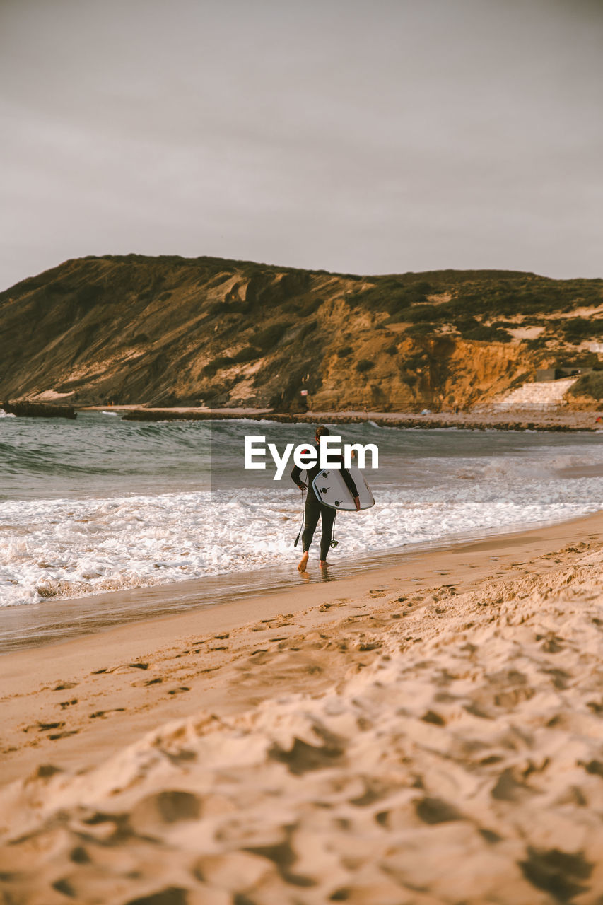 MAN WITH SURFBOARD WALKING AT BEACH AGAINST SKY