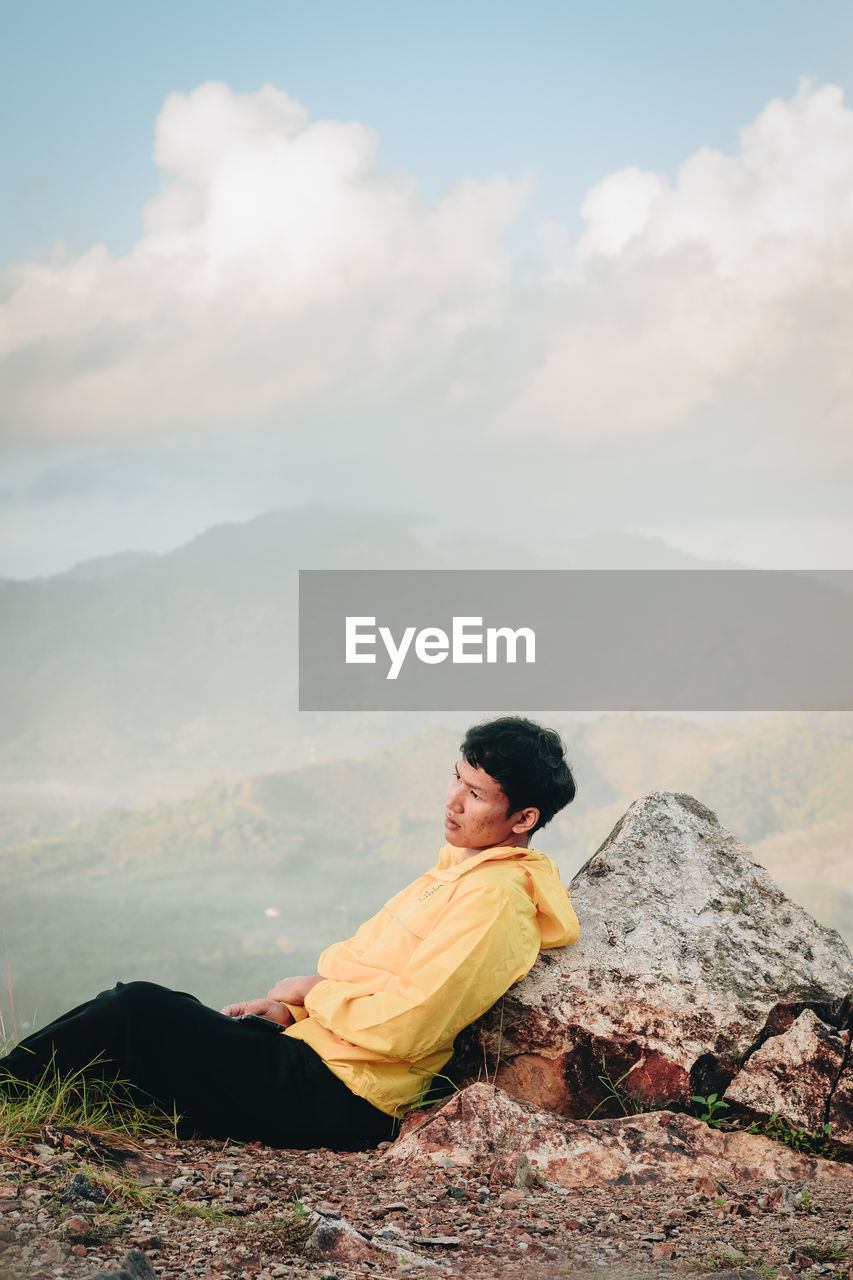 Side view of man sitting on mountain against cloudy sky