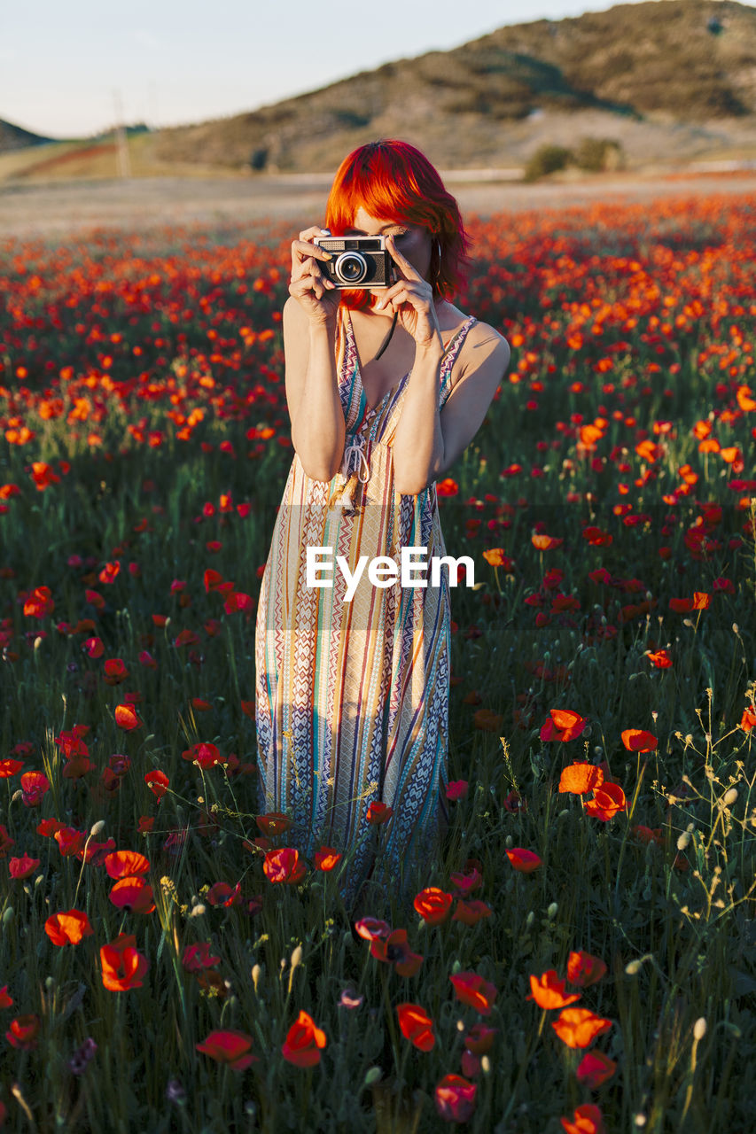 Redheaded woman photographing through camera while standing at poppy field