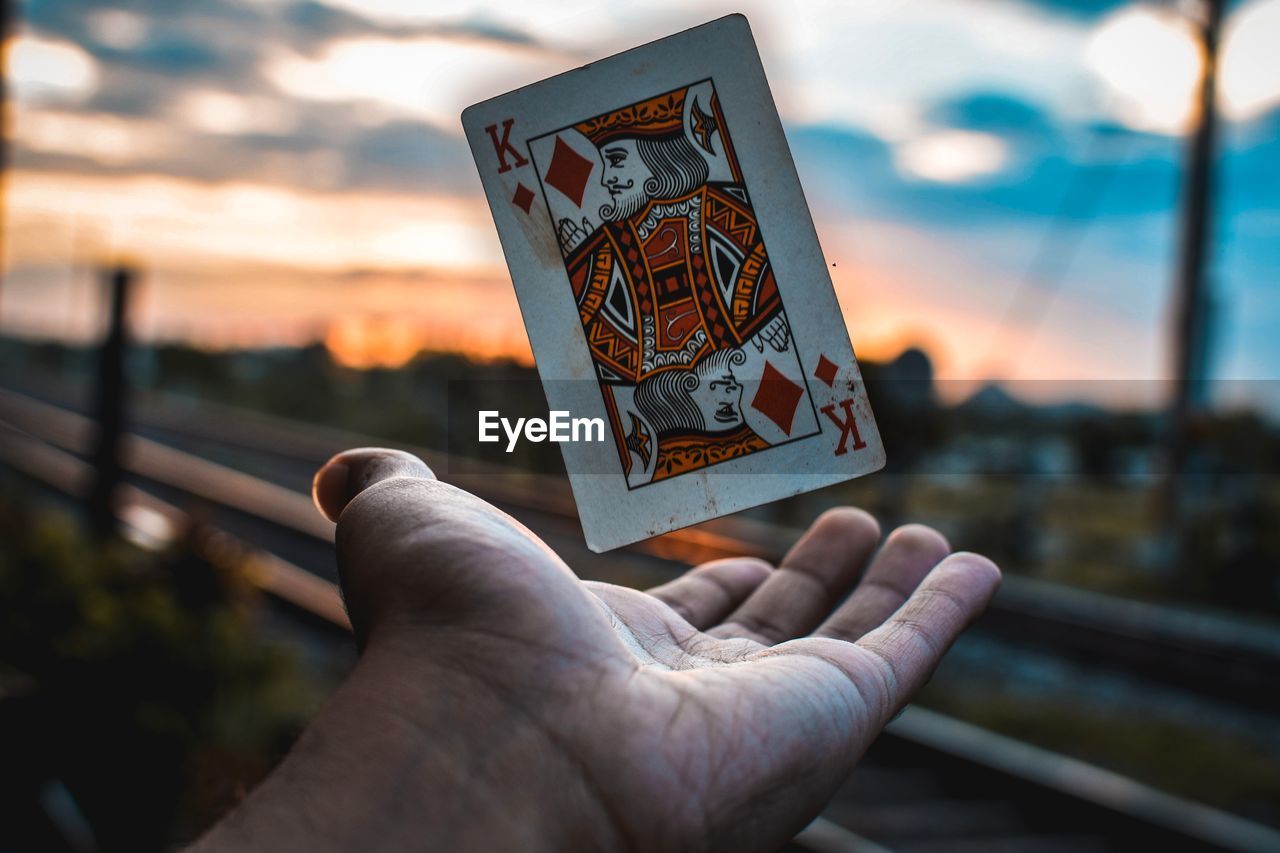 Close-up of hand catching king card against sky during sunset