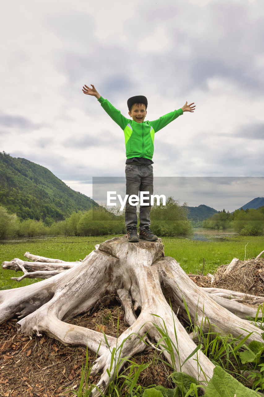 Full length of boy with arms outstretched standing on tree stump against cloudy sky