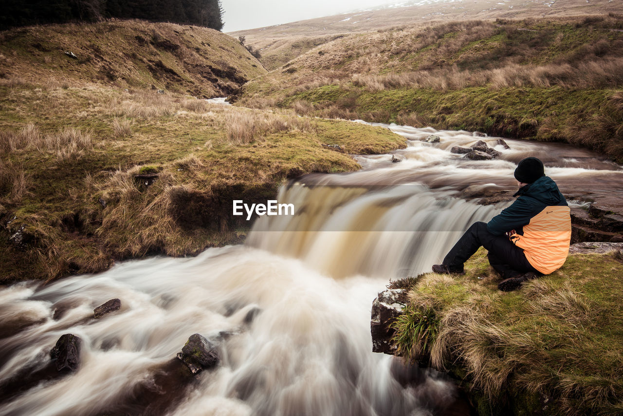 REAR VIEW OF MAN LOOKING AT WATERFALL AGAINST ROCKS