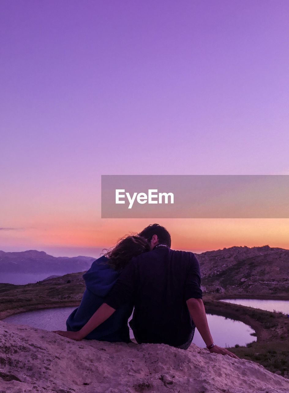 Rear view of couple romancing on rock by mountains against sky during sunset